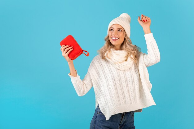 Mann, der einen drahtlosen Lautsprecher hält, der Musik hört und gerne tanzt, trägt einen weißen Pullover und eine Strickmütze im Winterstil, die isoliert auf Blau posiert