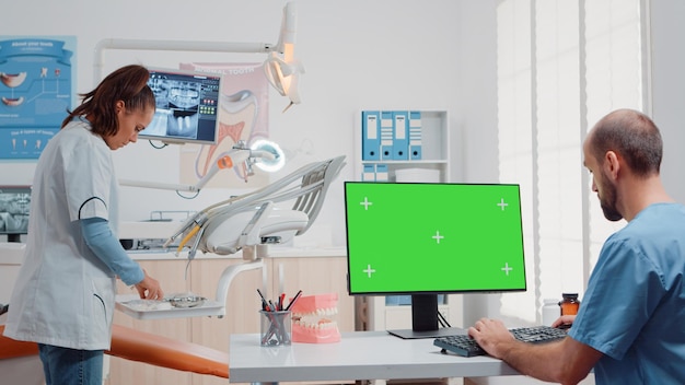 Mann, der einen Monitor mit horizontalem grünem Bildschirm verwendet und mit dem Zahnarzt über Mundpflege im Kabinett spricht. Spezialisten, die mit zahnärztlichen Werkzeugen für die Zahnpflege und isolierte Mockup-Vorlagen auf dem Computer arbeiten