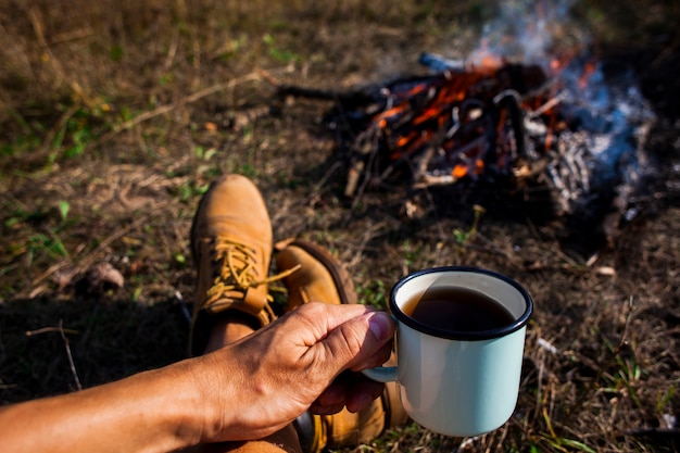 Mann, der einen Tasse Kaffee nahe bei einem Lagerfeuer hält