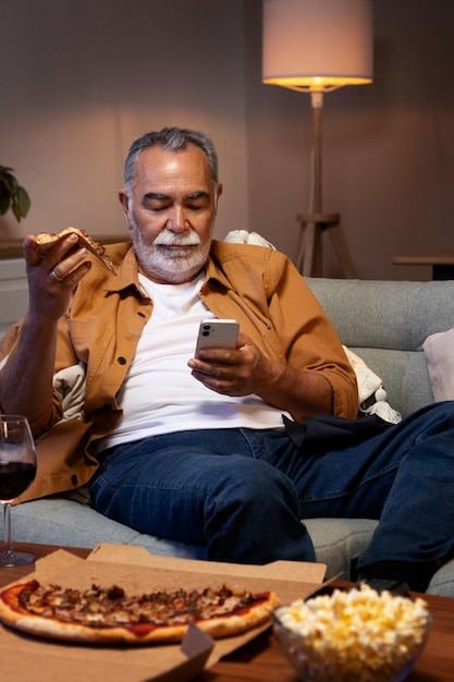 Mann, der etwas zu essen genießt, während er allein zu Hause ist und ein Smartphone benutzt