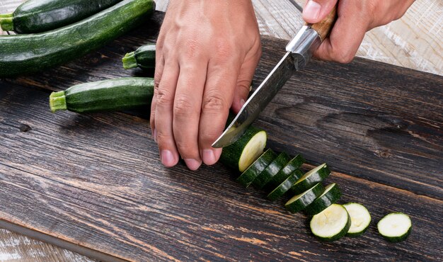 Mann, der frische Zucchini in Scheiben auf einem Schneidebrett auf einem Holztisch schneidet