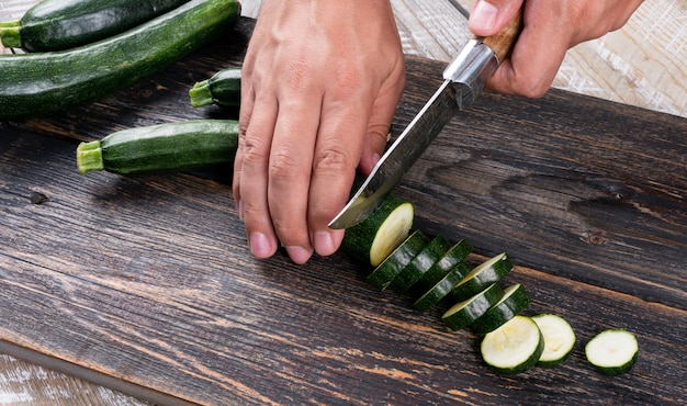 Mann, der frische Zucchini in Scheiben auf einem Schneidebrett auf einem Holztisch schneidet