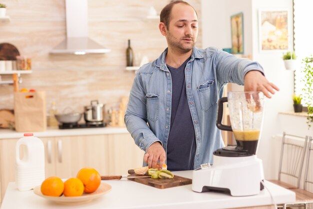 Mann, der Früchte in den Mixer legt, um sie für einen gesunden Smoothie zu mischen. Gesunder, unbeschwerter und fröhlicher Lebensstil, Ernährung und Frühstückszubereitung am gemütlichen sonnigen Morgen