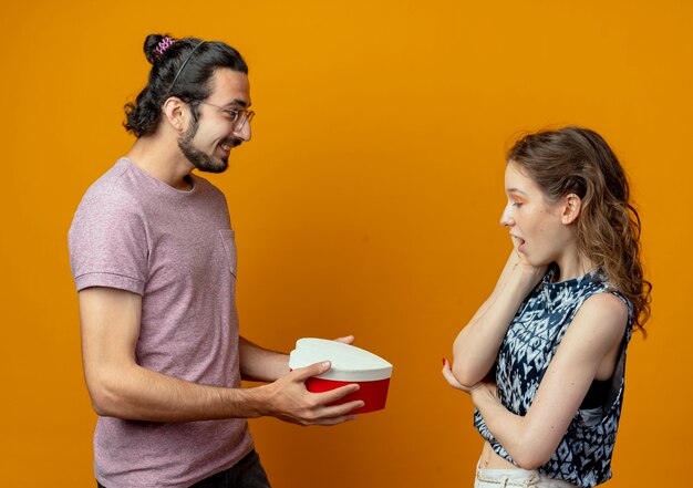 Mann, der Geschenkschachtel zu seiner Freundin, jungen schönen Paar Mann und Frauen über orange Hintergrund gibt