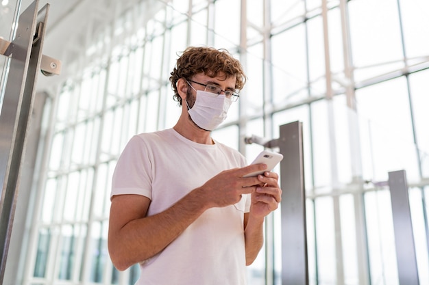 Mann, der Gesichtsmaske im Flughafen und im Smartphone trägt