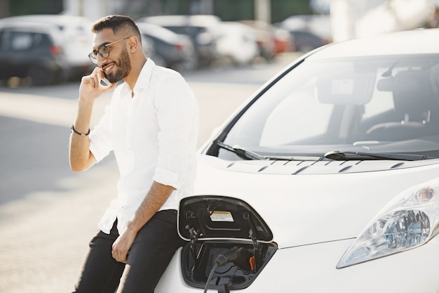 Mann, der mobiles gespräch hat, während er auf elektroauto wartet. ladestation, öko-autos. arabische ethnie.