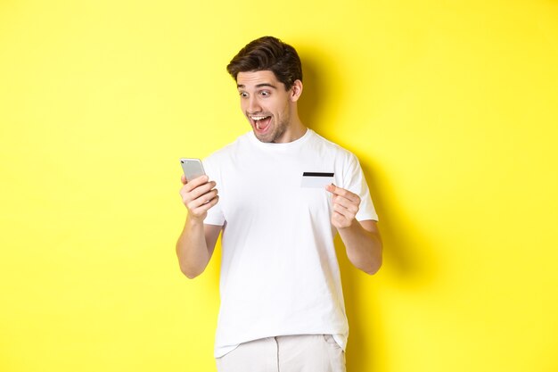 Mann, der überrascht am Smartphone schaut, online einkauft, Kreditkarte hält, über gelbem Hintergrund steht. Speicherplatz kopieren