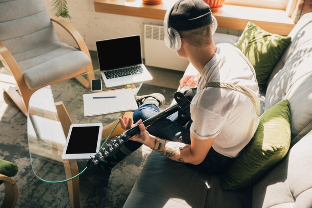 Mann, der während Online-Kursen zu Hause lernt oder sich selbst kostenlos informiert. Wird Musiker, Gitarrist, während er isoliert ist, Quarantäne gegen die Ausbreitung des Coronavirus. Mit Laptop, Smartphone, Kopfhörer.