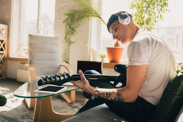 Mann, der während Online-Kursen zu Hause lernt oder sich selbst kostenlos informiert. Wird Musiker, Gitarrist, während er isoliert ist, Quarantäne gegen die Ausbreitung des Coronavirus. Mit Laptop, Smartphone, Kopfhörer.