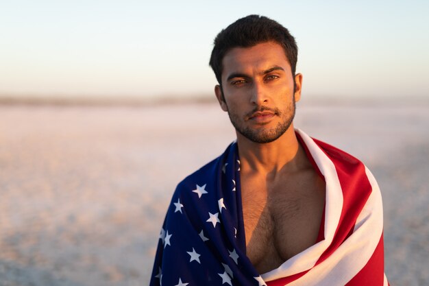 Mann eingewickelt in der amerikanischen Flagge, die auf dem Strand steht