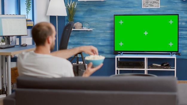 Mann entspannt sich auf der Couch und sieht sich im Fernsehen einen Film an, während er Popcorn im modernen Wohnzimmer isst. Person, die eine gute Zeit hat, auf dem Sofa zu sitzen und auf den Fernseher mit Chroma-Key-Display zu schauen.