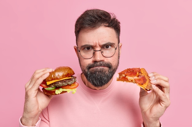 Kostenloses Foto mann hält hamburger und pizza presst lippen trägt einen runden brillenpullover