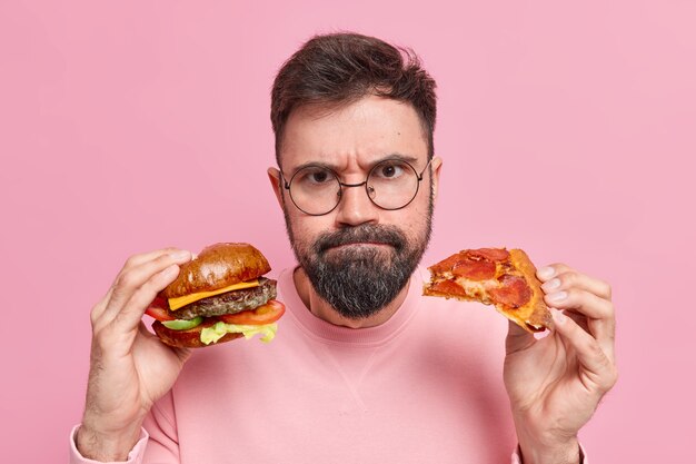 Mann hält Hamburger und Pizza presst Lippen trägt einen runden Brillenpullover