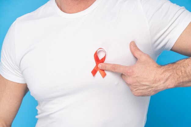 Mann hält rote Schleife AIDS-Aufklärungssymbol auf einem Herzen, das ein weißes T-Shirt trägt, das auf blauem Hintergrund isoliert ist Moderne Medizin und Gesundheitswesen AIDS-Aufklärungskonzept