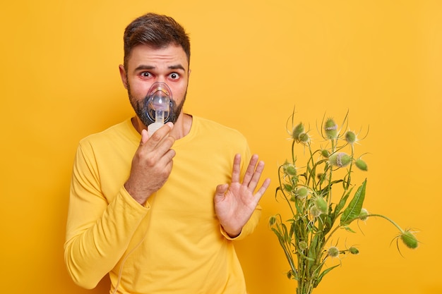 Mann hat Probleme mit der Lunge leidet an Asthma Allergiesymptome hat rote geschwollene Augen hält sich von Allergen fern, die gegen Pollen allergisch sind trägt Inhalationsmaske isoliert auf gelber Wand