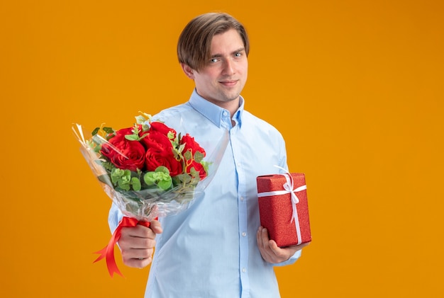 Mann im Blueshirt, der Strauß der roten Rosen hält und präsentierendes lächelndes selbstbewusstes Valentinstagkonzept über orange Wand steht