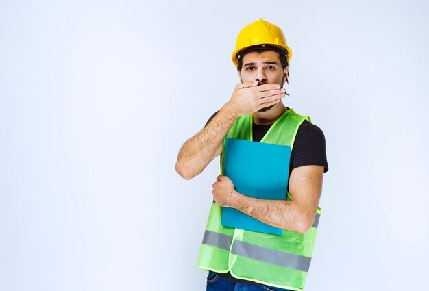 Mann im gelben Helm mit blauem Ordner sieht verängstigt und verängstigt aus.