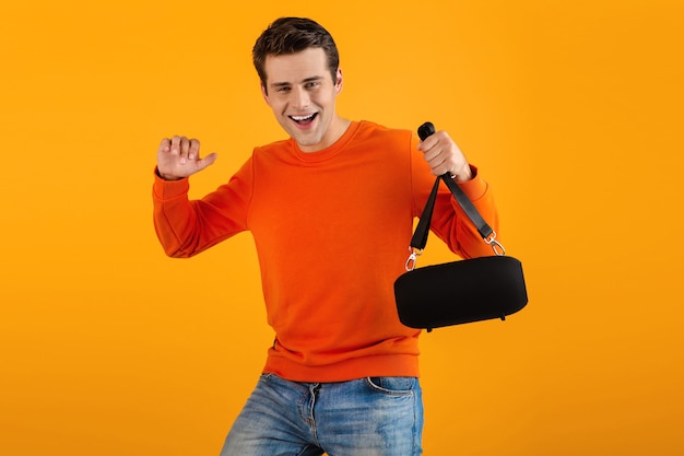 Mann im orangefarbenen pullover, der einen drahtlosen lautsprecher hält, der glücklich musik hört und spaß hat, isoliert auf gelb