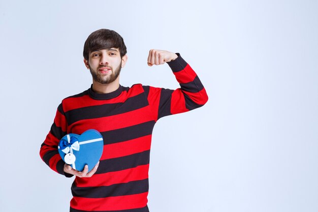 Mann im rot gestreiften Hemd, das eine blaue Herzform-Geschenkbox hält und seine Faust zeigt.