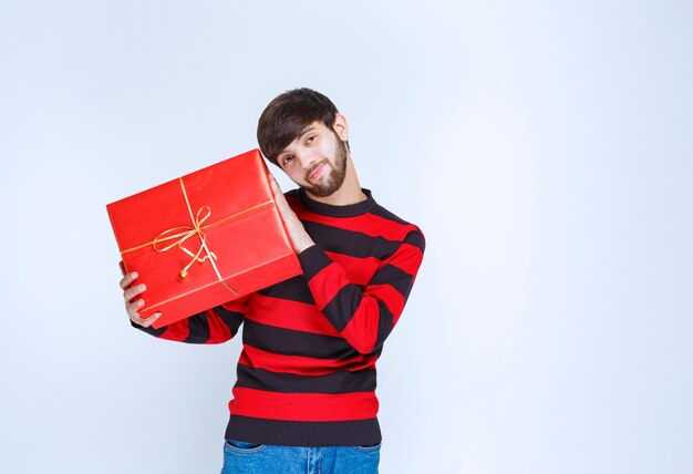 Mann im rot gestreiften Hemd, das eine rote Geschenkbox hält, sie dem Kunden oder seiner Freundin liefert und präsentiert. Foto in hoher Qualität