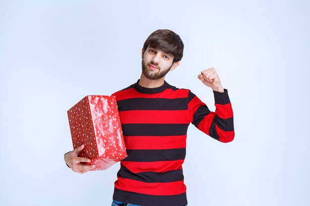 Mann im rot gestreiften Hemd, das eine rote Geschenkbox hält und sich kraftvoll und positiv fühlt.