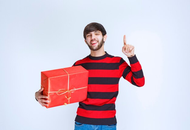 Mann im rot gestreiften Hemd, das eine rote Geschenkbox hält und verwirrt und nachdenklich aussieht.