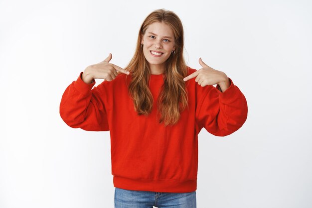 Mann im roten Pullover, der auf die Brust zeigt, wobei beide Zeigefinger freudig lächeln und dem Freund auf Grau das Outfit zeigen.