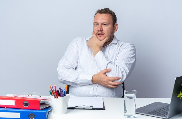 Mann im weißen Hemd, der mit nachdenklichem Ausdruck beiseite schaut, mit der Hand am Kinn am Tisch sitzend mit Laptop-Büroordnern und Zwischenablage über weißer Wand, die im Büro arbeitet