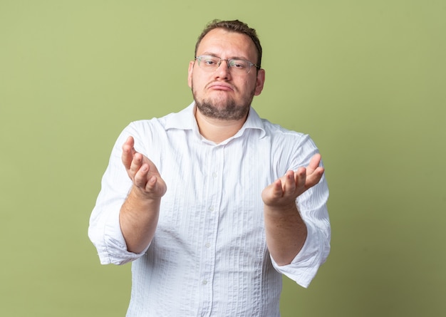 Mann im weißen Hemd mit Brille, der einen schiefen Mund mit enttäuschtem Gesichtsausdruck macht, der die Arme über der grünen Wand hebt