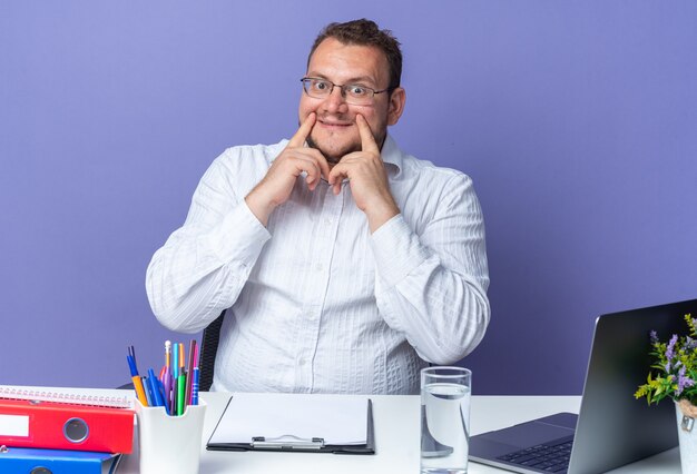 Mann im weißen Hemd mit Brille, der mit Zeigefingern auf sein falsches Lächeln zeigt, das am Tisch mit Laptop und Büroordnern über blauer Wand im Büro sitzt