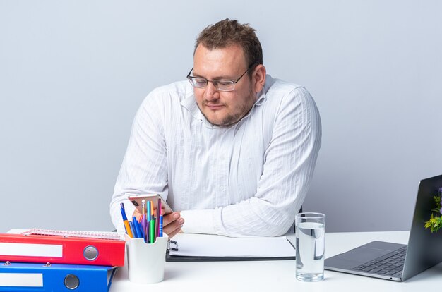 Mann im weißen Hemd mit Brille, der müde und gelangweilt am Tisch sitzt, mit Laptop-Büroordnern und Zwischenablage über weißer Wand im Büro arbeitet