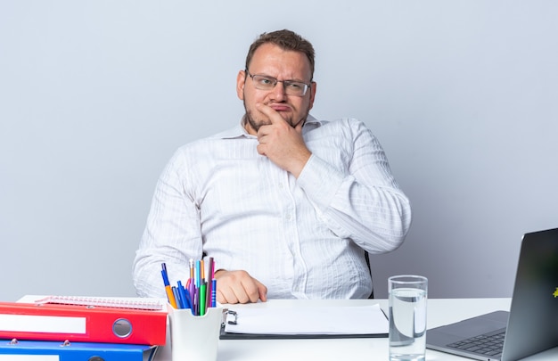 Mann im weißen Hemd mit Brille, der verwirrt am Tisch sitzt, mit Laptop-Büroordnern und Zwischenablage auf weißem Hintergrund, der im Büro arbeitet