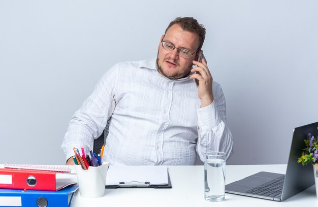 Mann im weißen Hemd mit Brille telefoniert mit ernstem Gesicht am Tisch mit Laptop-Büroordnern und Zwischenablage auf Weiß