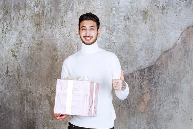 Mann im weißen Pullover, der eine lila Geschenkbox hält, die mit einem Band umwickelt ist und ein erfolgreiches Handzeichen zeigt.