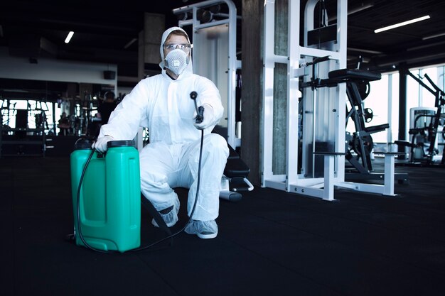 Mann im weißen Schutzanzug desinfiziert und Fitnessgeräte und Gewichte, um die Ausbreitung des hoch ansteckenden Koronavirus zu stoppen