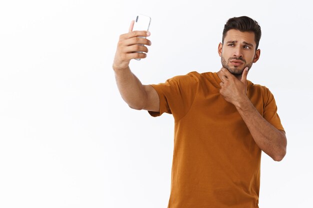 Mann in braunem T-Shirt, wie er aussieht, Selfie mit Smartphone in erhobener Hand machen, Bart berühren und kokett die Lippen falten