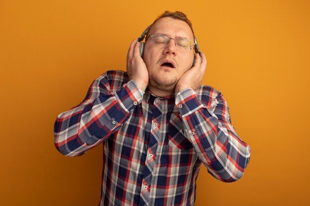 Mann in Brille und kariertem Hemd mit Kopfhörern, die Musik genießen, die über orange Wand stehen