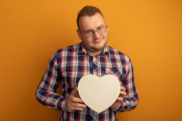 Kostenloses Foto mann in den gläsern und im karierten hemd, das cardboradherz hält, das zuversichtlich über orange wand steht