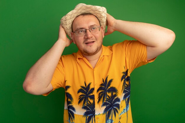 Mann in der Brille, die orange Hemd und Sommerhut trägt, die mit glücklichem Gesicht mit hnads hinter seinem Kopf stehen, der über grüner Wand steht