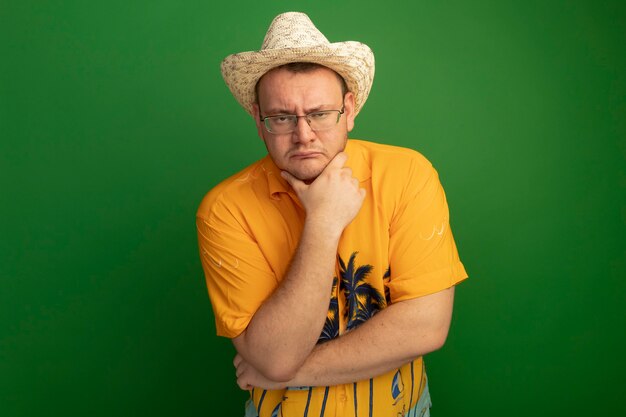 Mann in der Brille, die oranges Hemd und Sommerhut mit Hand auf seinem Kinn trägt und mit ernstem Gesicht über grüner Wand steht