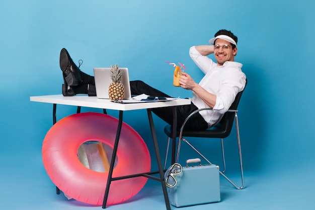 Mann in der Mütze arbeitet im Urlaub und trinkt Cocktail. Guy sitzt mit Koffer, aufblasbarem Kreis, Laptop und Ananas am Tisch.
