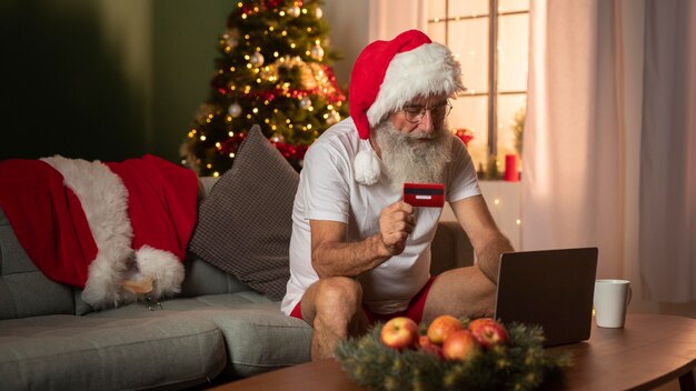 Mann in der Weihnachtsmütze, die Kreditkarte hält und online mit Laptop zu Hause einkauft