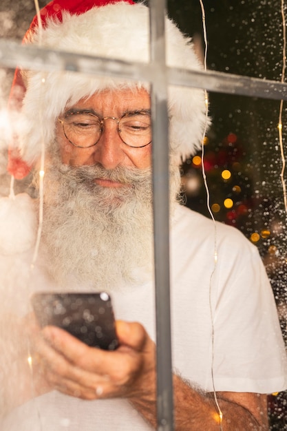 Mann in der Weihnachtsmütze unter Verwendung des Smartphones durch Fenster