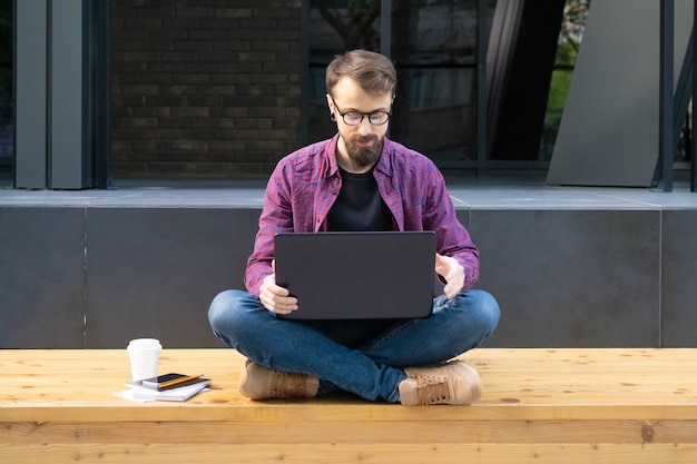Mann in Gläsern, die mit gekreuzten Beinen auf Holzbank mit Laptop sitzen