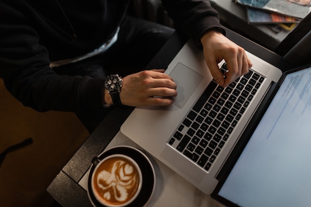 Mann in schwarzer kleidung mit einer luxusuhr arbeitet an einem laptop und trinkt kaffee in einem café. typ mit computer und cappuccino