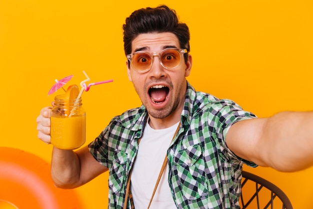 Mann in Sonnenbrille und grünem Hemd macht ein Selfie und schaut überrascht in die Kamera. Porträt des Kerls mit Glas Cocktail auf isoliertem Raum.
