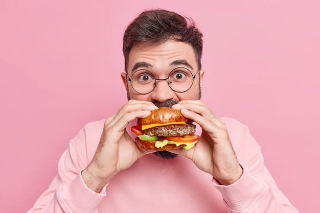 Mann isst gierig leckeren Hamburger hat großen Hunger konsumiert Fast Food trägt runde Brille und Pullover