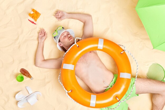 Mann ist am Strand eingeschlafen liegt auf warmem weißem Sand mit Rettungsring auf dem Bauch genießt den Sommerreiseurlaub hat einen faulen Tag umgeben von Hausschuhen Sonnenschirm erfrischendes Getränk Tennisschläger