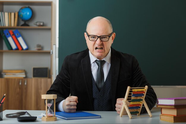 Mann, Lehrer, der eine Brille mit Klassenbuch trägt, sitzt an der Schulbank vor der Tafel im Klassenzimmer und benutzt Abakus, der die Fäuste ballt und mit aggressivem Ausdruck schreit