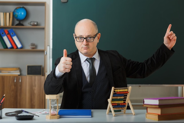 Mann, Lehrer, der eine Brille mit Klassenbuch trägt, sitzt an der Schulbank vor der Tafel im Klassenzimmer und sieht selbstbewusst aus, zeigt den Daumen nach oben und zeigt mit dem Zeigefinger nach oben